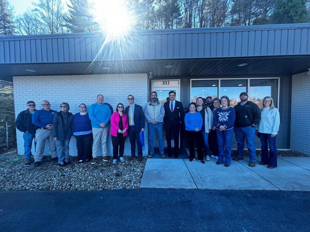 A group of people stands in a line outside a building under bright sunlight, with trees and a blue sky in the background. The group includes men and women in casual and formal clothing.