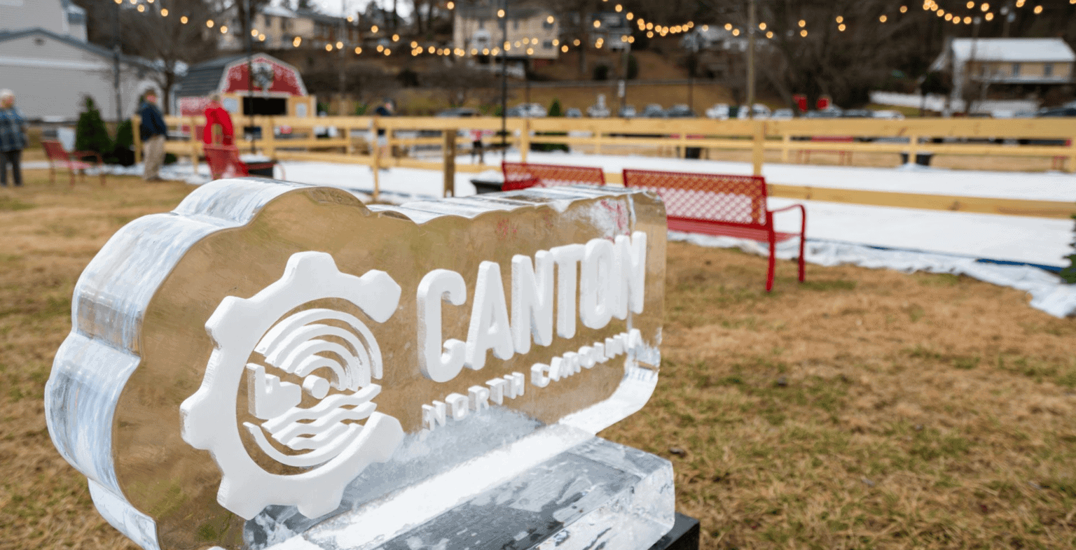 A carved ice sculpture with Canton North Carolina stands in front of an outdoor ice skating rink, with string lights and people in the background on a grassy area.