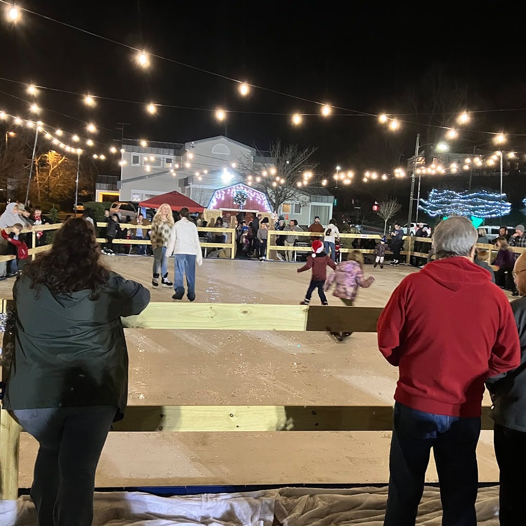 People watch children ice skating at night under string lights. A crowd stands around a fenced rink. Festive lights and decorations adorn the background buildings, creating a lively winter scene.