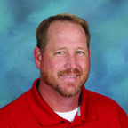 A man with short light brown hair, a beard, and a mustache smiles at the camera. He is wearing a red collared shirt, in front of a blue studio backdrop.