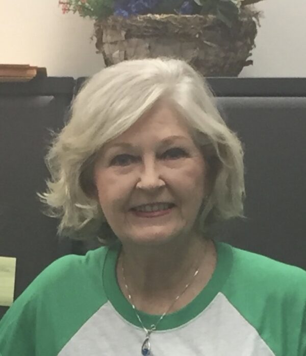 A woman with short, wavy gray hair smiles at the camera. She is wearing a green and white top and a necklace. There is a plant with flowers in the background.