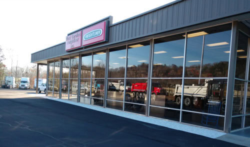 A modern commercial building with large glass windows reflects trucks and trees outside. The building has a gray exterior and a sign above the entrance. The parking lot in front is empty and newly paved.