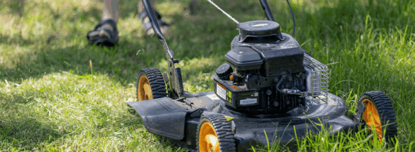 A person wearing sandals pushes a black lawn mower with orange wheels across green grass on a sunny day.