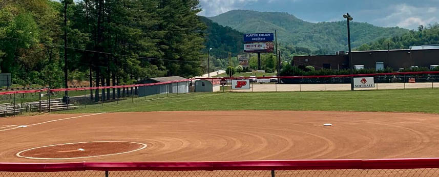 A softball field with bases, a red fence, bleachers on the left, and mountains in the background under a partly cloudy sky. A scoreboard and buildings are visible beyond the outfield.
