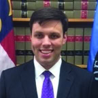 A man in a suit and purple tie smiles in front of a bookshelf filled with law books, with two flags visible on either side.